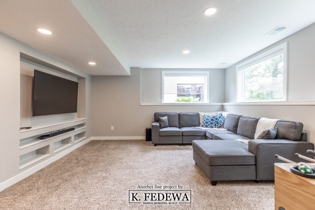 Holt, Michigan basement remodel from K. Fedewa Builders with a couch, wall-mounted tv, tan carpet, and bright sun coming in through two windows.