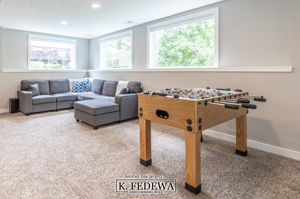 Gaming area in Holt, Michigan basement remodel from K. Fedewa Builders with a couch and foosball table, while sunlight pours in through three windows.