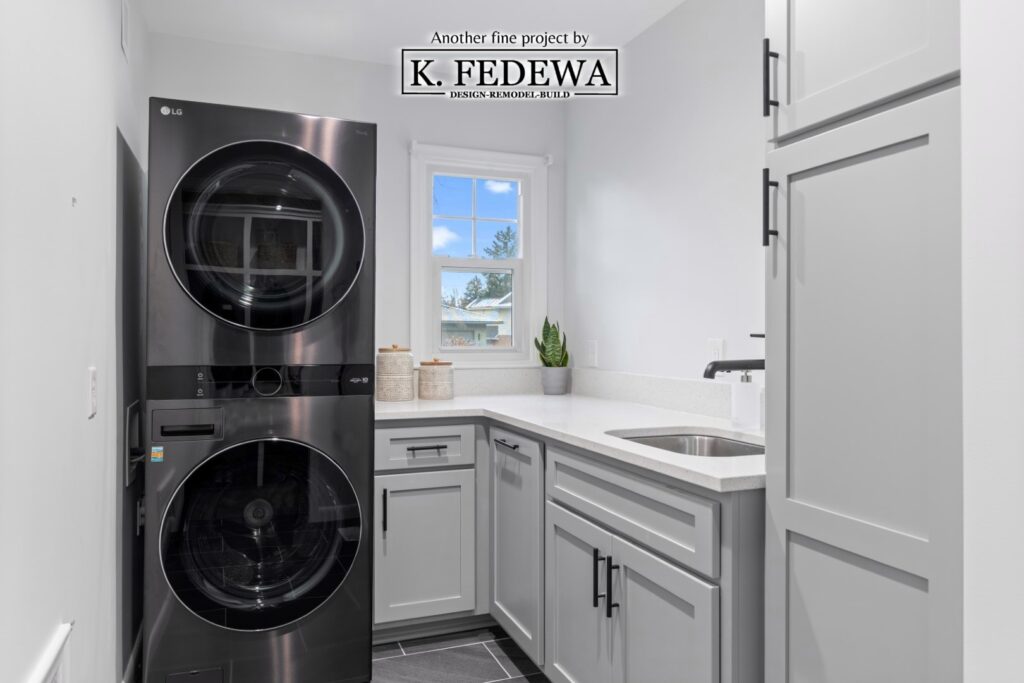 Remodeled laundry room with black stacked washer and dryer, along with a lot of grey cabinets with white countertops.