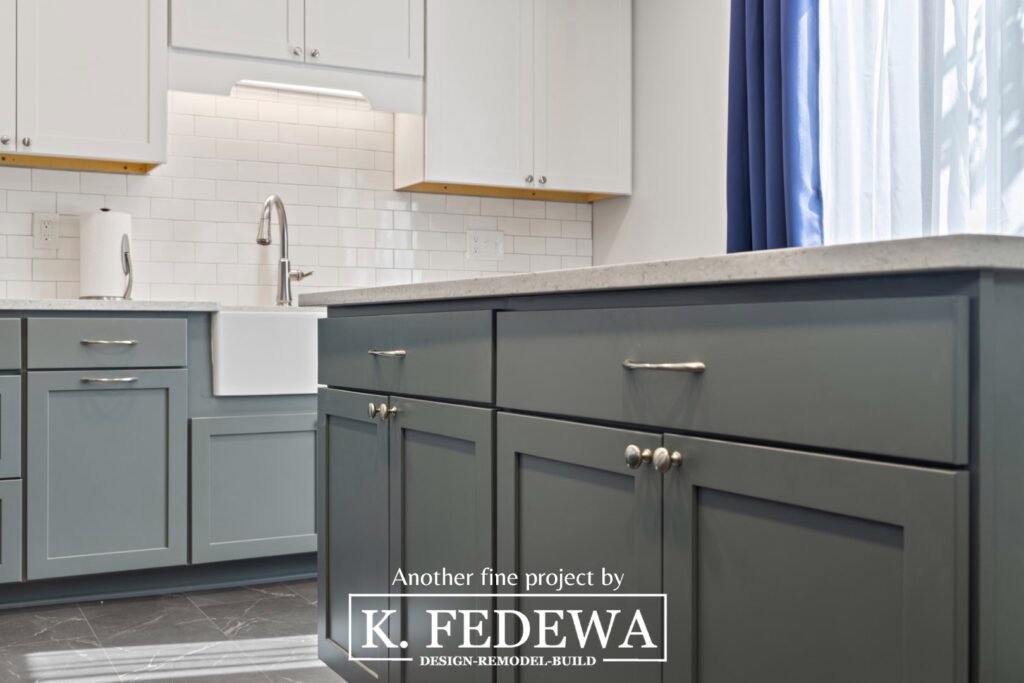 Close-up of sage green cabinets in a kitchen island with a white apron-style sink, sage green lower cabinets, white subway tile backsplash, and white upper cabinets in the background.