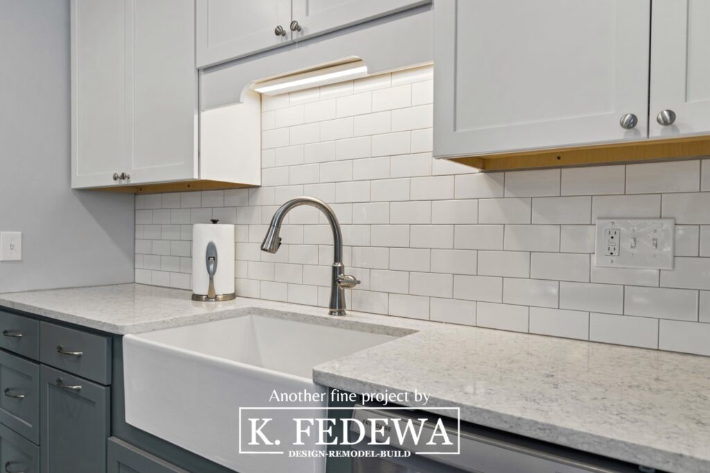 Close-up of apron style sink on top of blue cabinets, white quartz countertops, and below a white subway tile backsplash and white upper cabinets.